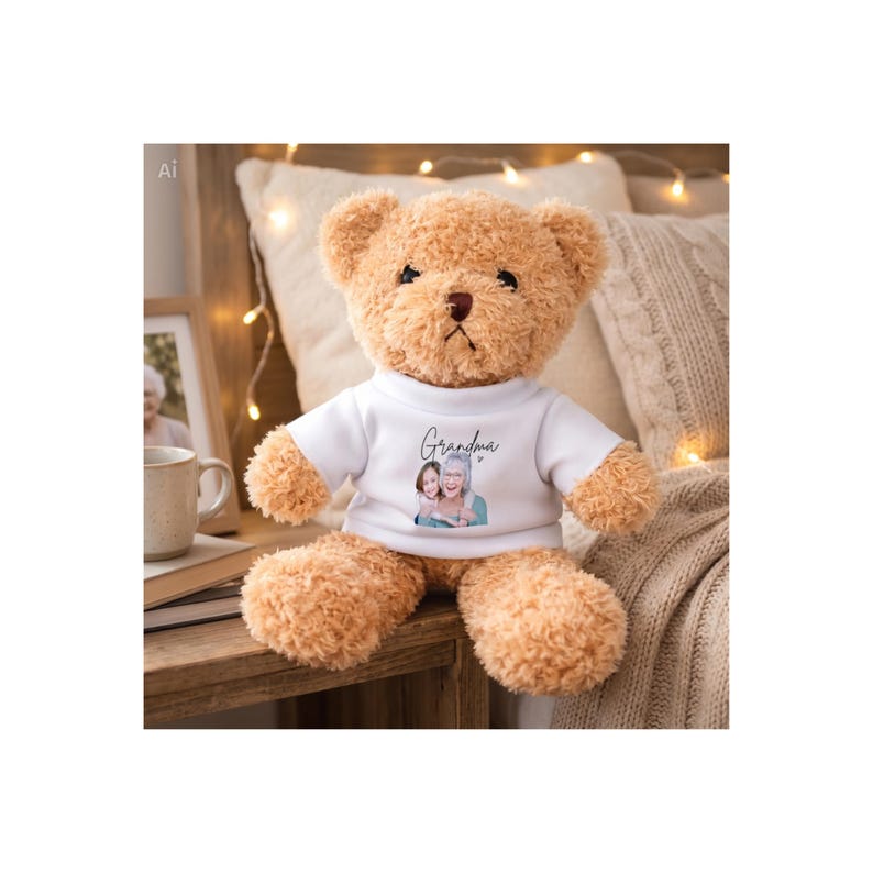 Op de afbeelding: Een zachte, lichtbruine teddybeer met een wit t-shirt. Het t-shirt toont het woord "Oma" in script boven een afbeelding van een vrouw en een meisje. De beer heeft een bruine neus en zit op een houten oppervlak.