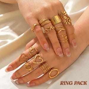 Puede incluir: Una colección de anillos dorados con varios diseños, incluyendo formas de corazón, bandas y motivos de hojas. Los anillos se muestran en una mano con uñas largas y cuidadas. El texto "RING PACK" es visible en la parte inferior.
