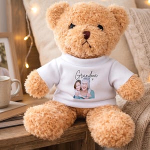 Op de afbeelding: Een lichtbruine teddybeer met een wit t-shirt met het woord "Oma" en een afbeelding van een vrouw en een kind. De beer is gemaakt van pluche materiaal en zit op een houten oppervlak.