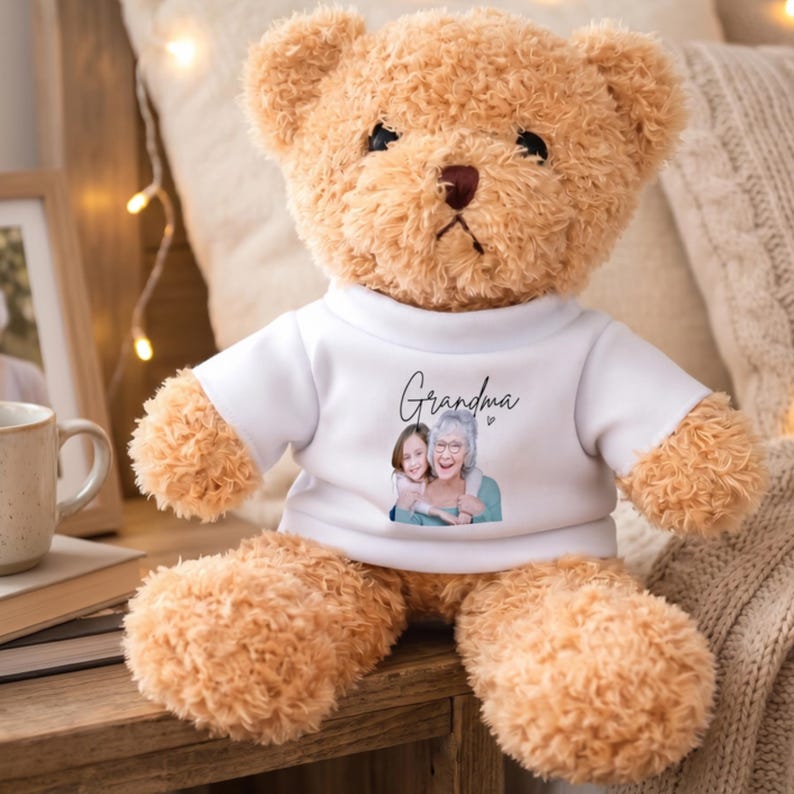 Op de afbeelding: Een lichtbruine teddybeer met een wit t-shirt met het woord "Oma" en een afbeelding van een vrouw en een kind. De beer is gemaakt van pluche materiaal en zit op een houten oppervlak.