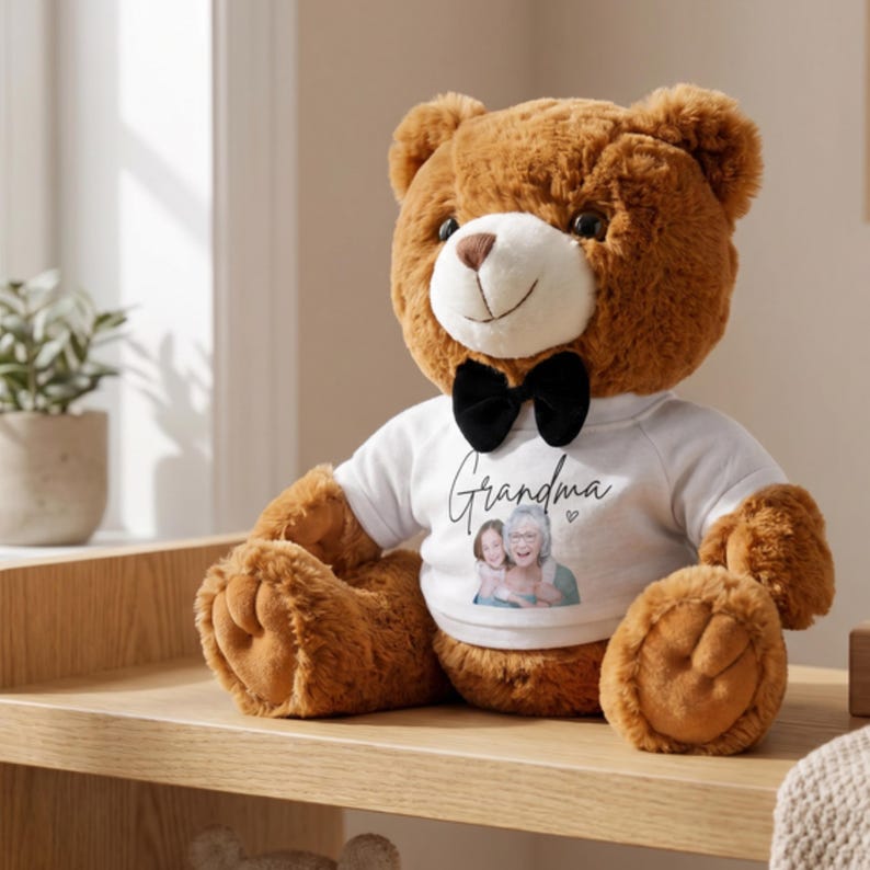 Op de afbeelding: Een bruine teddybeer met een witte snuit en een zwarte vlinderdas. De beer draagt een wit T-shirt met het woord "Oma" en een foto van twee personen. De beer zit op een houten plank.