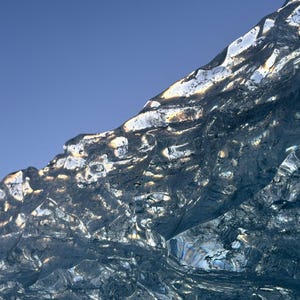 Op de afbeelding: Een close-up van een gletsjer met een diepblauwe kleur. Het ijsoppervlak is getextureerd met verschillende vormen en patronen, die zonlicht reflecteren. De bovenrand van de gletsjer is verlicht, wat een contrast creëert met de heldere blauwe lucht.