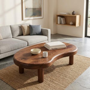 May include: A kidney-shaped coffee table made of dark brown wood with a smooth, polished surface and four thick, cylindrical legs. On the table sits a small cream-colored bowl, a white cylindrical cup, and two stacked books with the word Textiles.