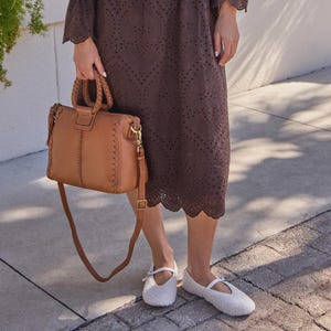May include: A brown leather handbag with a braided handle and a detachable shoulder strap. The bag has decorative stitching and gold-colored hardware. The person is wearing a brown dress and white shoes.