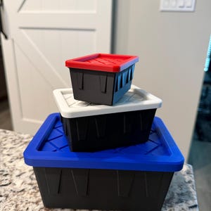 May include: Three black storage bins with colored lids stacked on top of each other. The top bin has a red lid, the middle bin has a white lid, and the bottom bin has a blue lid. The bins are sitting on a countertop.
