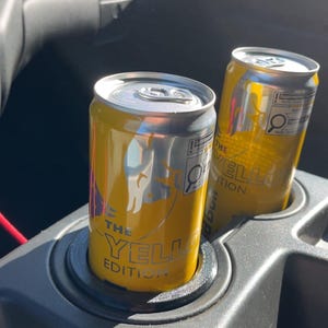 May include: Two yellow and silver cans labeled "THE YELLOW EDITION" are in a car's cup holder. The cans have a silver top and a yellow body with a logo and text. The cup holder is black.