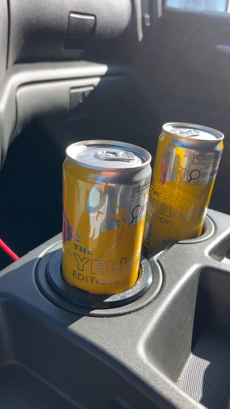 May include: Two yellow and silver cans labeled "THE YELLOW EDITION" are in a car's cup holder. The cans have a silver top and a yellow body with a logo and text. The cup holder is black.