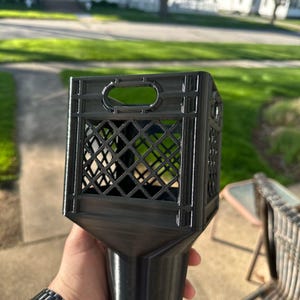 May include: A black, 3D-printed lantern with a square top and a lattice design. The lantern has a handle and a tapered base. The image is taken outdoors, with a blurred background of a house and greenery.