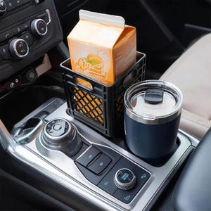 May include: A black plastic crate holding an orange juice carton sits in a car's console. A navy blue insulated tumbler with a silver lid is next to the crate. The car's gear shift and control panel are visible.