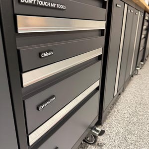May include: A matte black metal tool chest with silver handles and multiple drawers. Labels on the drawers read DON'T TOUCH MY TOOLS, Chisels, Extensions, and Allen Wrenches. The unit is mounted on wheels and features a wooden top surface.