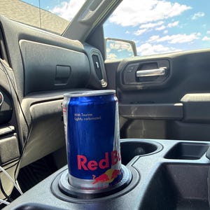 May include: A blue Red Bull energy drink can in a car's cup holder. The can has the Red Bull logo and the words "With Taurine. Lightly carbonated."