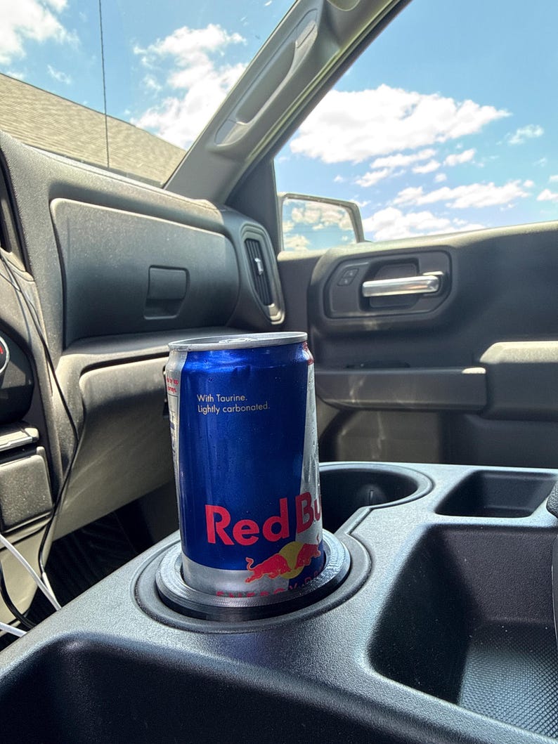 May include: A blue Red Bull energy drink can in a car's cup holder. The can has the Red Bull logo and the words "With Taurine. Lightly carbonated."