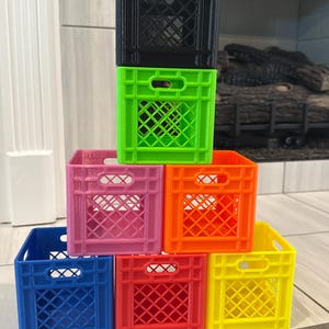 May include: A stack of seven miniature milk crates in various colors: black, green, pink, orange, blue, red, and yellow. Each crate has a rectangular shape with a grid pattern and handle cutouts, creating a colorful display.