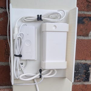 May include: A white plastic wall-mounted cable management bracket holding a white rectangular power adapter and a smaller white control module with a power button. White electrical cords are neatly coiled and secured with black zip ties.