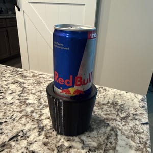 May include: A blue and silver Red Bull energy drink can inside a black 3D-printed cup holder. The can has the Red Bull logo and text. The cup holder is on a speckled countertop.