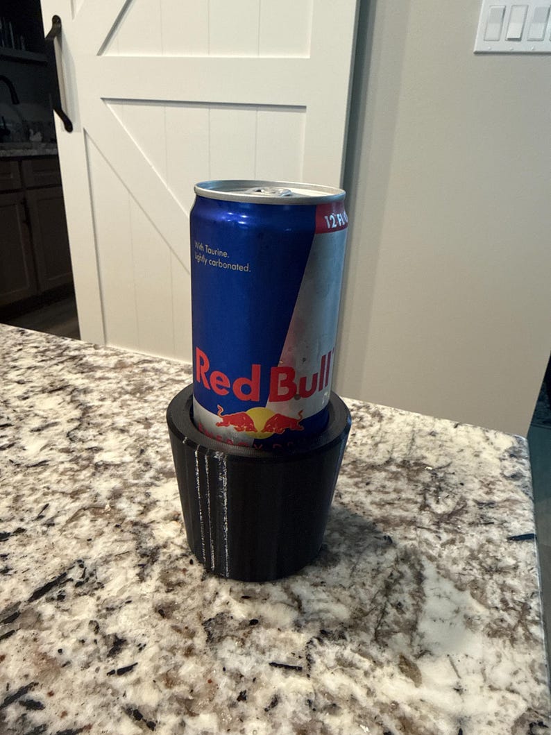 May include: A blue and silver Red Bull energy drink can inside a black 3D-printed cup holder. The can has the Red Bull logo and text. The cup holder is on a speckled countertop.