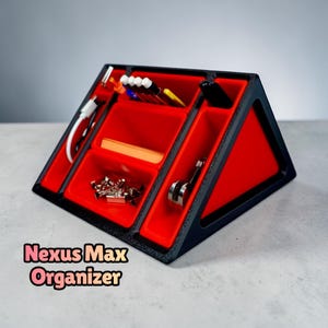 May include: A black triangular desk organizer with red interior compartments, labeled Nexus Max Organizer. It features multiple slots holding pens, a white charging cable, metal binder clips, and small office supplies in a structured layout.