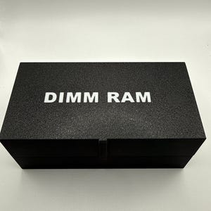 May include: A black rectangular box with the text "DIMM RAM" in white capital letters. The box has a textured surface and a simple, minimalist design. The box is likely used for storing or packaging computer components.