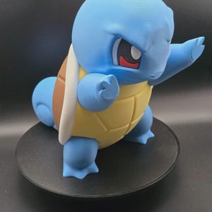 May include: A blue Squirtle figurine with a yellow and brown shell. The figurine is posed with its arms out, and is standing on a black circular base. The eyes are white with red accents.