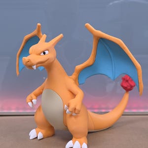 Könnte beinhalten: Orangefarbene Charizard-Figur mit blauen Flügeln und einer roten Flamme am Schwanz. Die Figur hat weiße Krallen und einen beigen Bauch. Der Drache steht mit den Armen an den Seiten.