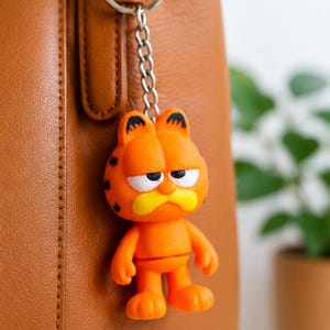 Peut inclure: Porte-clés Garfield orange avec des taches noires et une moustache jaune. Le porte-clés est attaché à une chaîne et un anneau argentés. Le fond est un sac en cuir marron.