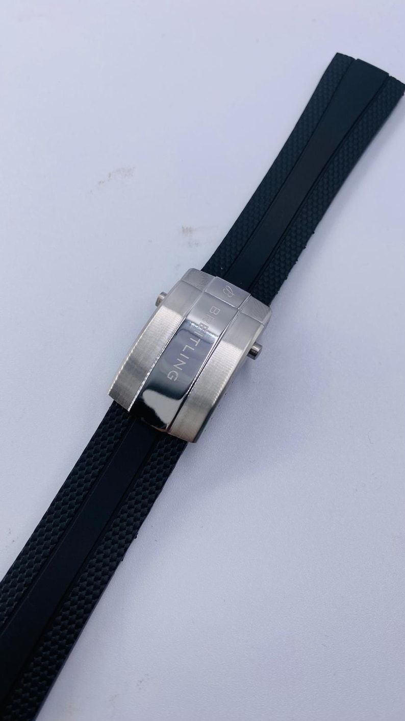 K&ouml;nnte beinhalten: Ein schwarzes Kautschuk-Uhrenarmband mit strukturierter Oberfl&auml;che und einer silberfarbenen Metall-Faltschlie&szlig;e. Die Schlie&szlig;e zeigt das eingravierte Breitling-Logo und den Markennamen auf ihrer geb&uuml;rsteten und polierten Oberfl&auml;che.