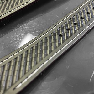 May include: Close-up of two watch straps. The straps are dark gray with a textured, woven pattern. White stitching runs along the edges. The straps have multiple holes for adjustment and a metal clasp.