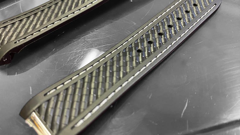 May include: Close-up of two watch straps. The straps are dark gray with a textured, woven pattern. White stitching runs along the edges. The straps have multiple holes for adjustment and a metal clasp.
