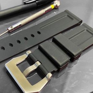 May include: Black watch strap with a silver buckle, alongside a small screwdriver and a pen. The strap has several holes for adjustment. The items are on a reflective surface.