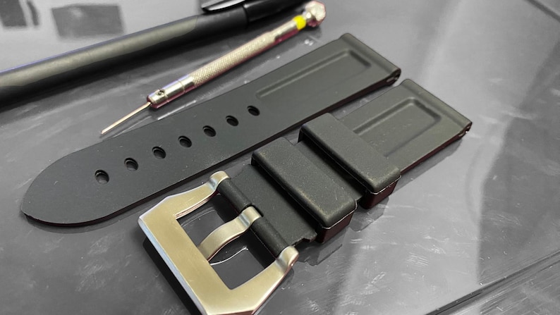 May include: Black watch strap with a silver buckle, alongside a small screwdriver and a pen. The strap has several holes for adjustment. The items are on a reflective surface.