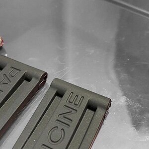 May include: Close-up of two black watch straps with the brand name "PANERAI" embossed on them. A small silver and yellow screwdriver and a black tool are in the background. The straps are made of a flexible material.