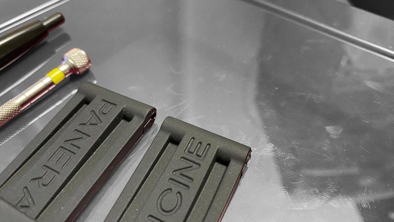 May include: Close-up of two black watch straps with the brand name "PANERAI" embossed on them. A small silver and yellow screwdriver and a black tool are in the background. The straps are made of a flexible material.