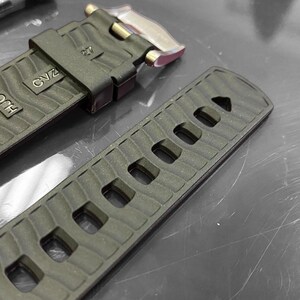May include: Close-up of a dark green watch strap with a textured wave pattern. The strap has several rectangular holes and is marked with the numbers "20" and "27", along with the letters "HUG" and "OV2". A silver clasp is visible.