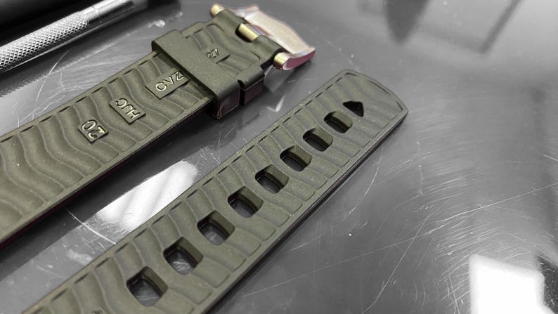 May include: Close-up of a dark green watch strap with a textured wave pattern. The strap has several rectangular holes and is marked with the numbers "20" and "27", along with the letters "HUG" and "OV2". A silver clasp is visible.