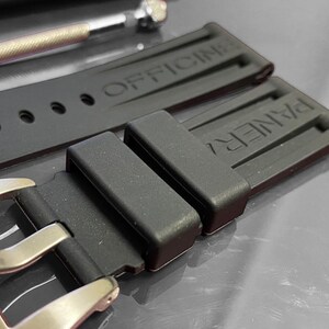 May include: Close-up of a black watch strap with the brand name "PANERAI" visible on the buckle and strap. The strap is made of a flexible material and has the word "OFFICINE" embossed on it. A small metal tool is in the background.
