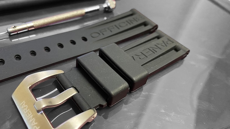 May include: Close-up of a black watch strap with the brand name "PANERAI" visible on the buckle and strap. The strap is made of a flexible material and has the word "OFFICINE" embossed on it. A small metal tool is in the background.