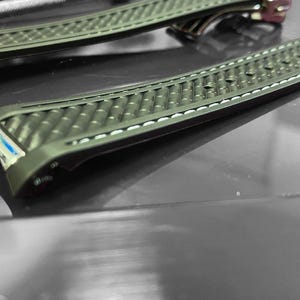 May include: Two olive green watch straps with a textured pattern and white stitching. The straps have a curved design and are made of a durable material. The watch straps are displayed on a reflective surface.