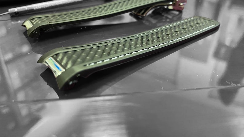 May include: Two olive green watch straps with a textured pattern and white stitching. The straps have a curved design and are made of a durable material. The watch straps are displayed on a reflective surface.