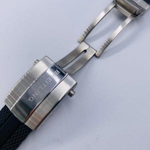 K&ouml;nnte beinhalten: Eine silberfarbene Uhren-Faltschlie&szlig;e aus Metall, befestigt an einem schwarzen, strukturierten Kautschuk-Uhrenarmband. Die Schlie&szlig;e hat eine geb&uuml;rstete Oberfl&auml;che und den eingravierten Schriftzug BREITLING auf dem Mittelteil sowie einen Druckknopf.