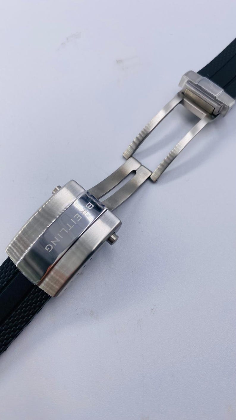 K&ouml;nnte beinhalten: Eine silberfarbene Uhren-Faltschlie&szlig;e aus Metall, befestigt an einem schwarzen, strukturierten Kautschuk-Uhrenarmband. Die Schlie&szlig;e hat eine geb&uuml;rstete Oberfl&auml;che und den eingravierten Schriftzug BREITLING auf dem Mittelteil sowie einen Druckknopf.