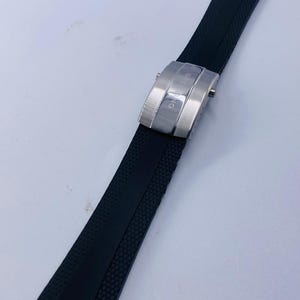 May include: A black rubber watch strap featuring a textured surface and a silver-toned metal deployment clasp. The clasp is engraved with the text BREITLING and includes two side release buttons for securing the strap around a wrist.
