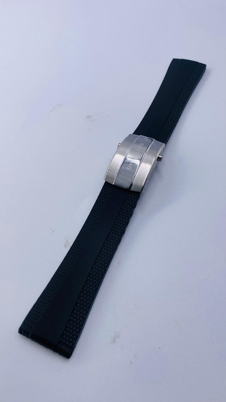 May include: A black rubber watch strap featuring a textured surface and a silver-toned metal deployment clasp. The clasp is engraved with the text BREITLING and includes two side release buttons for securing the strap around a wrist.