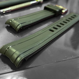 May include: Two olive green watch straps with a textured design. One strap has a gold-colored buckle. A black pen and a silver pen are in the background. The straps are on a reflective surface.