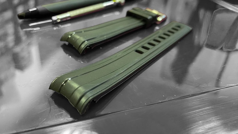 May include: Two olive green watch straps with a textured design. One strap has a gold-colored buckle. A black pen and a silver pen are in the background. The straps are on a reflective surface.