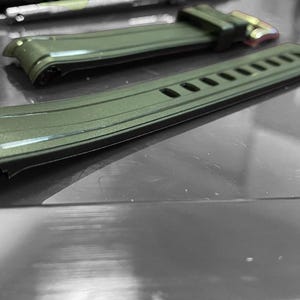 May include: Close-up of two olive green watch straps. The straps are made of a flexible material with a textured surface. One strap has a buckle with gold-colored metal. The other strap has several rectangular cutouts.