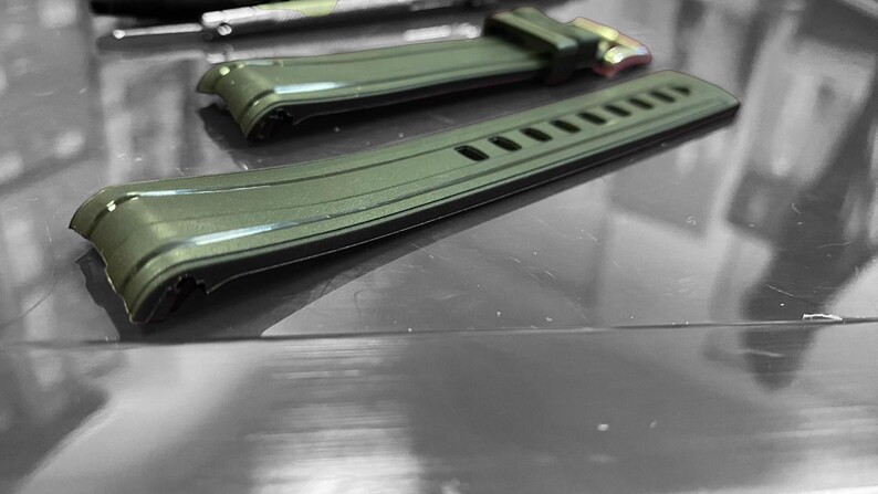 May include: Close-up of two olive green watch straps. The straps are made of a flexible material with a textured surface. One strap has a buckle with gold-colored metal. The other strap has several rectangular cutouts.
