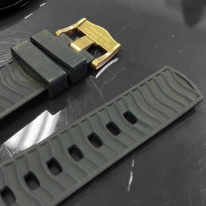 May include: Close-up of a dark gray watch strap with a gold-colored buckle. The strap has a textured wave pattern and several holes for adjustment. The buckle is engraved with text.