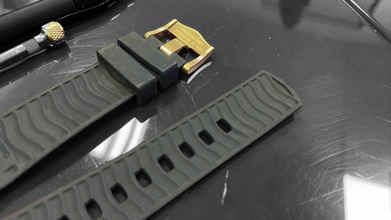 May include: Close-up of a dark gray watch strap with a gold-colored buckle. The strap has a textured wave pattern and several holes for adjustment. The buckle is engraved with text.
