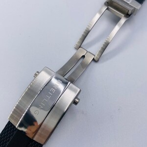 May include: A black rubber watch strap featuring a silver-toned stainless steel deployment clasp. The metal buckle is engraved with the word BREITLING in capital letters. The strap has a textured surface and the clasp is shown in an open position.