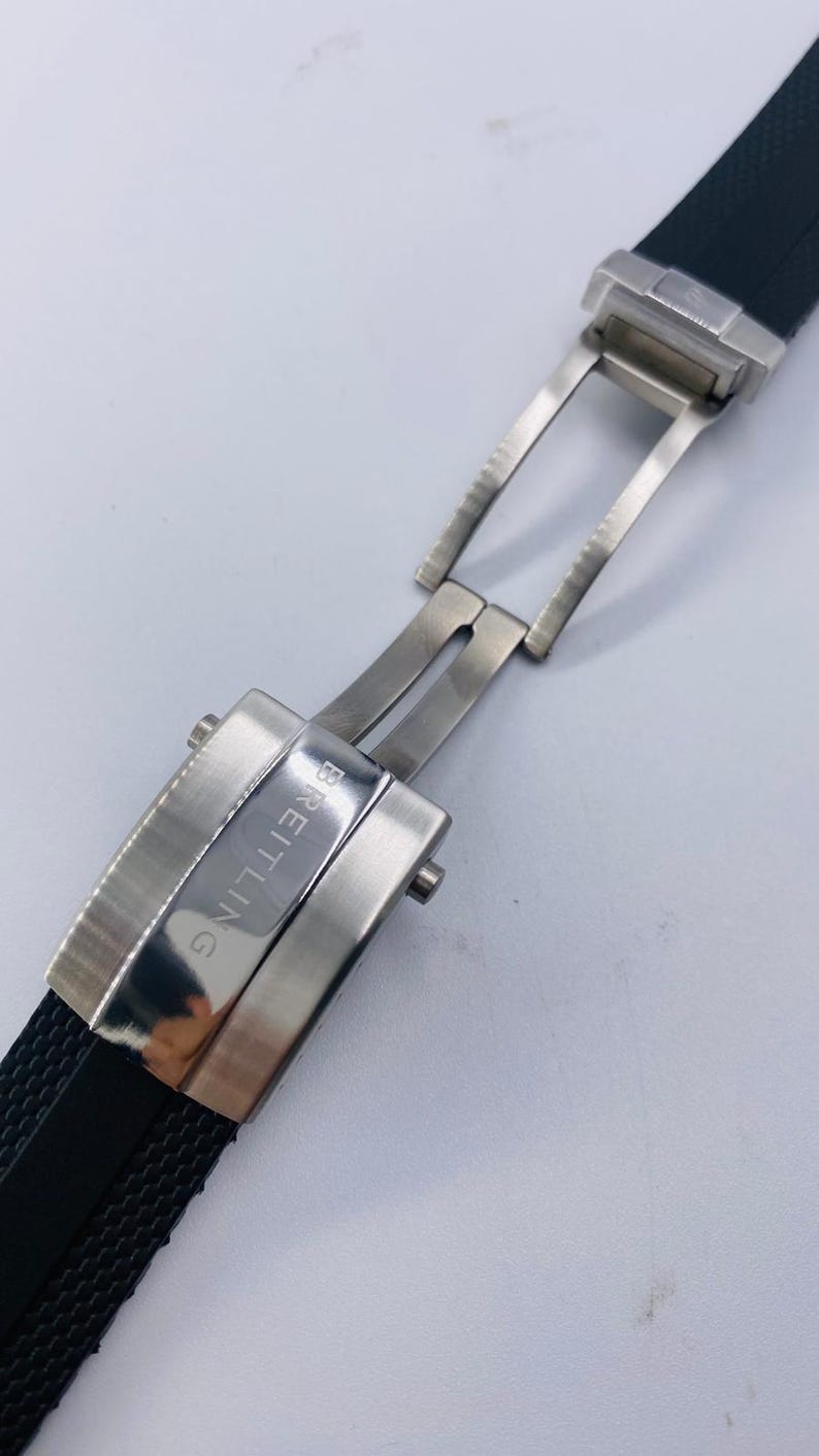May include: A black rubber watch strap featuring a silver-toned stainless steel deployment clasp. The metal buckle is engraved with the word BREITLING in capital letters. The strap has a textured surface and the clasp is shown in an open position.